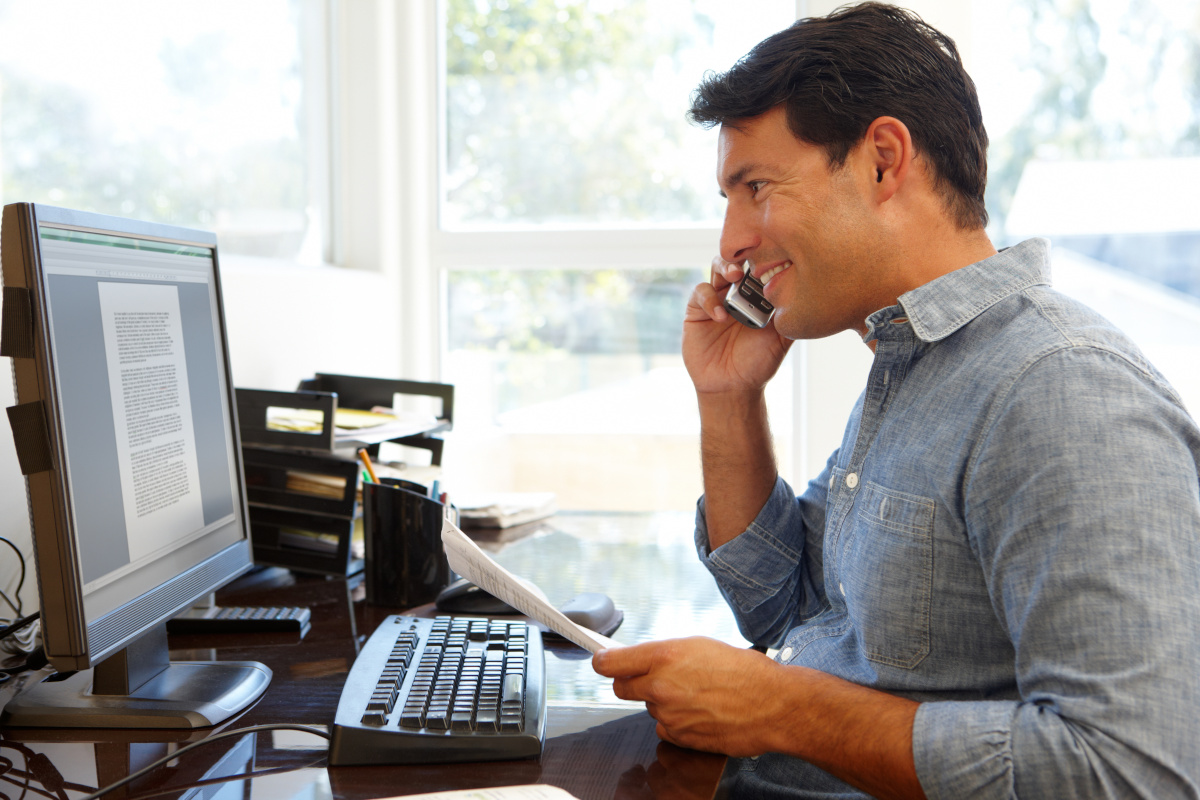 Smiling man holding papers while talking on phone and looking at computer